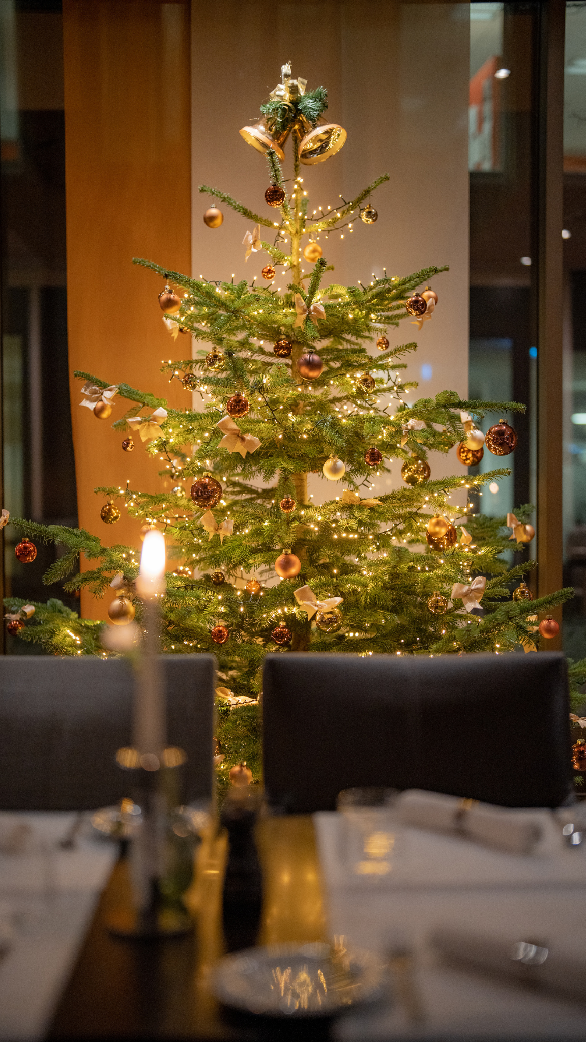 Geschmückter, beleuchteter Weihnachtsbaum in dunklem Restaurant, im Vordergrund unscharf ein Tisch mit Stühlen und brennender Kerze